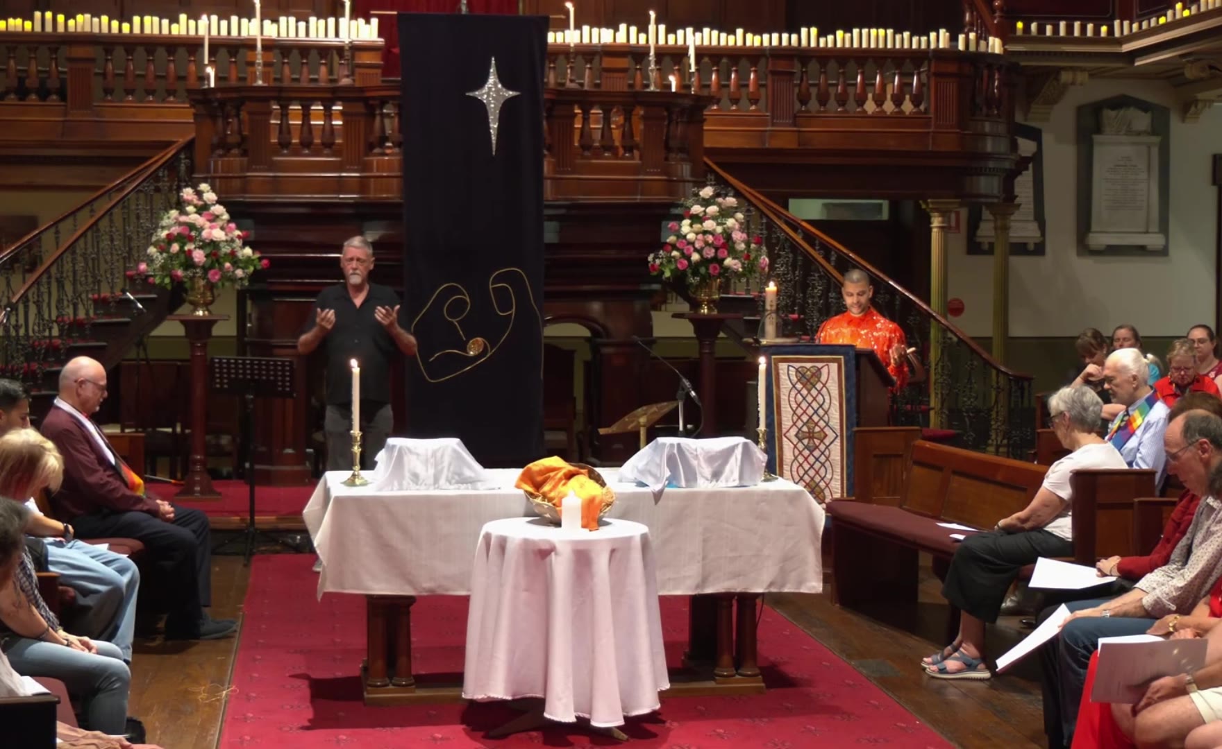 A wide view of the church: candles along the gallery rail, a dark banner between the stairs, the congregation seated on both sides, and Traidy Naidoo at the lectern on the right in an orange fringed shirt, hands open as he speaks.