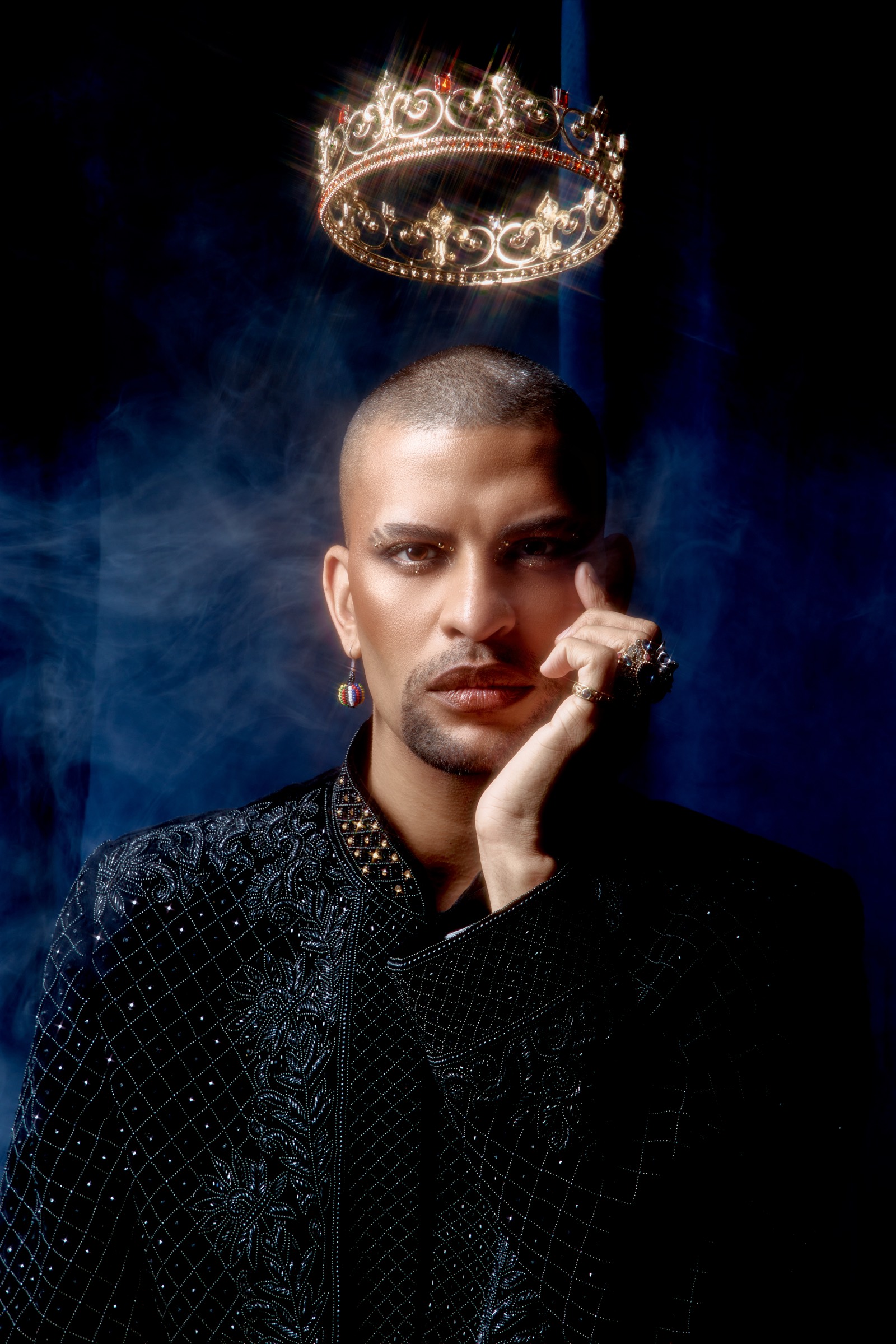 Traidy Naidoo in a black beaded jacket beneath a floating crown, in low light.