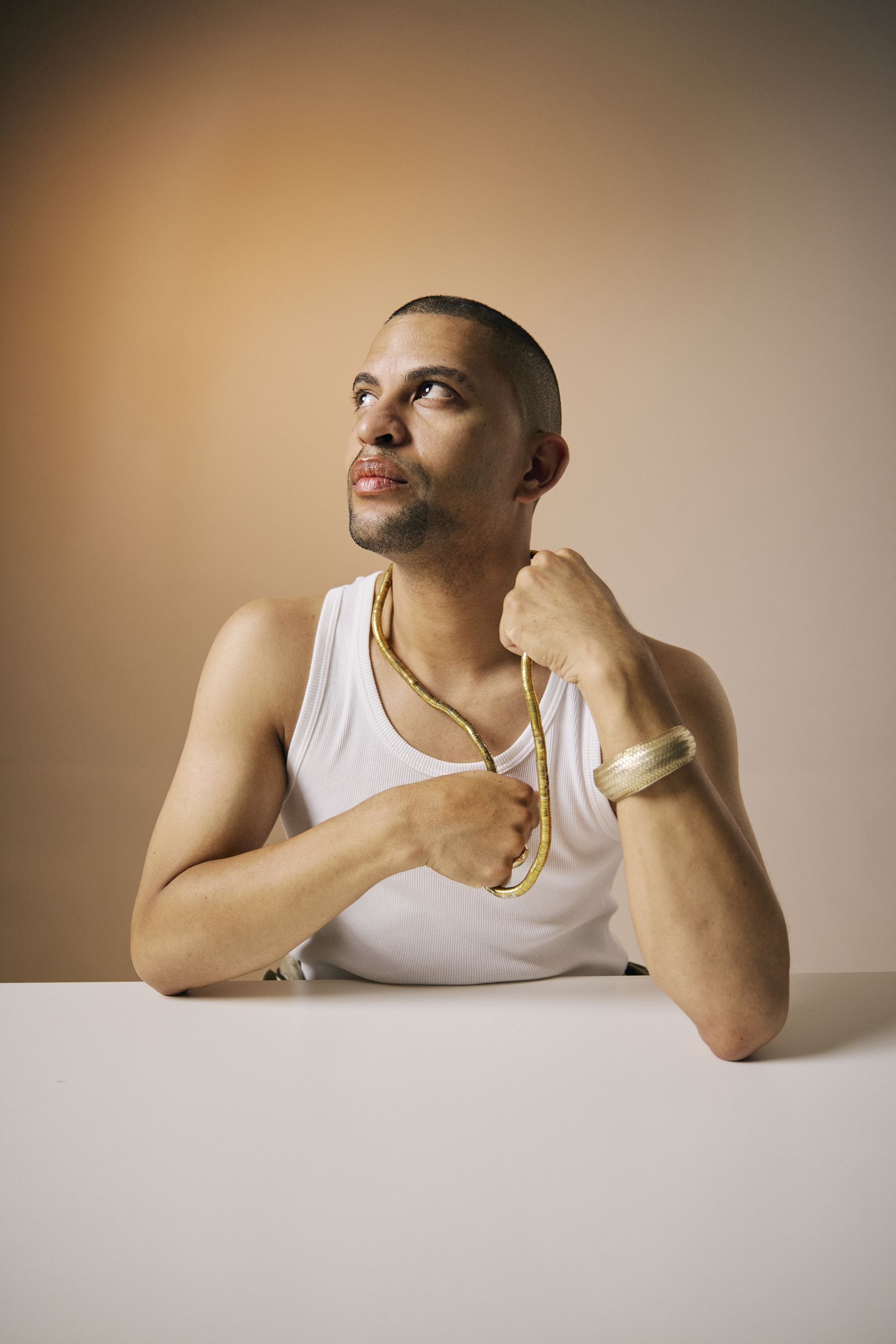 Traidy Naidoo in a white vest and gold jewellery against an apricot backdrop, looking up.