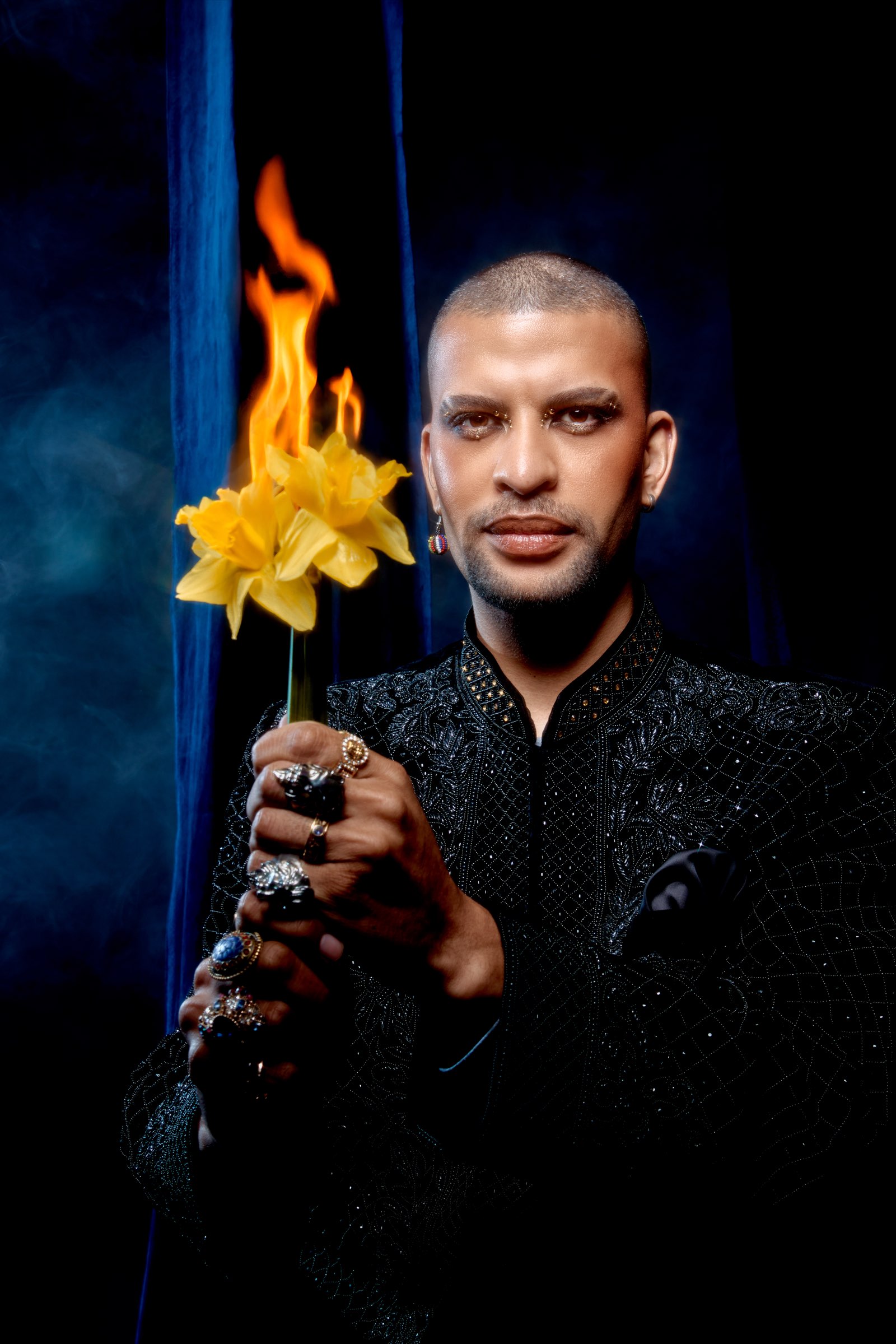 Traidy Naidoo in a black beaded jacket holding a burning stem of daffodils, blue velvet behind him.