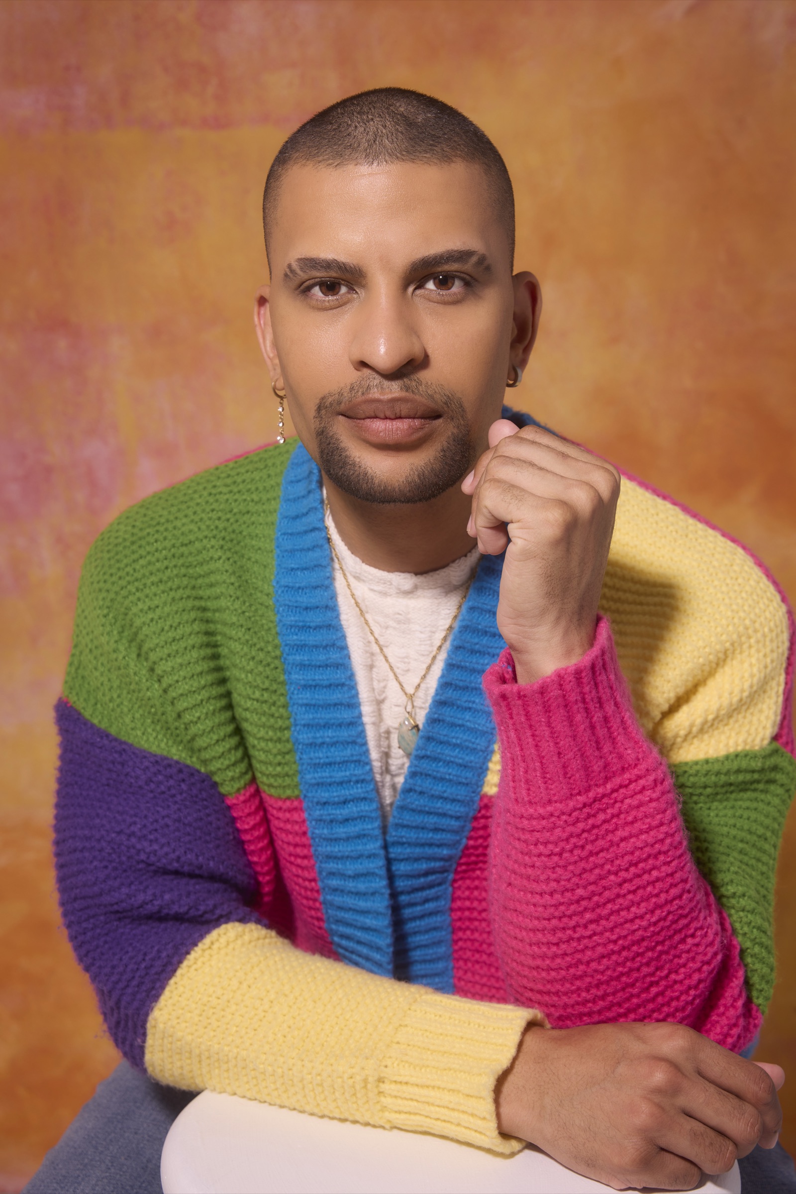 Traidy Naidoo seated in a colour-blocked knit cardigan, hand to his chin.