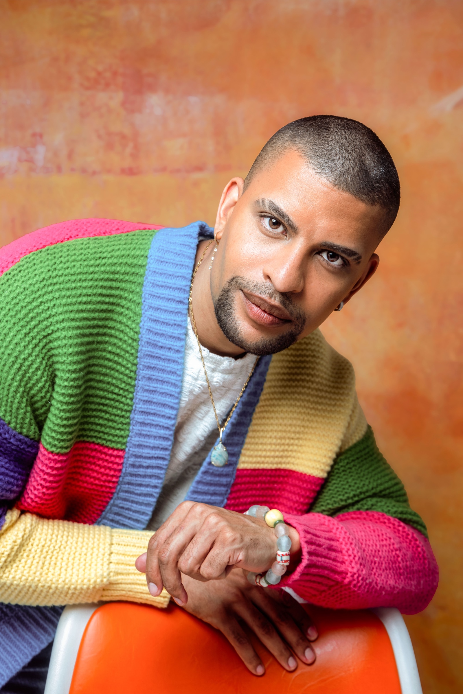 Traidy Naidoo leaning on an orange stool in a colour-blocked knit cardigan, smiling.