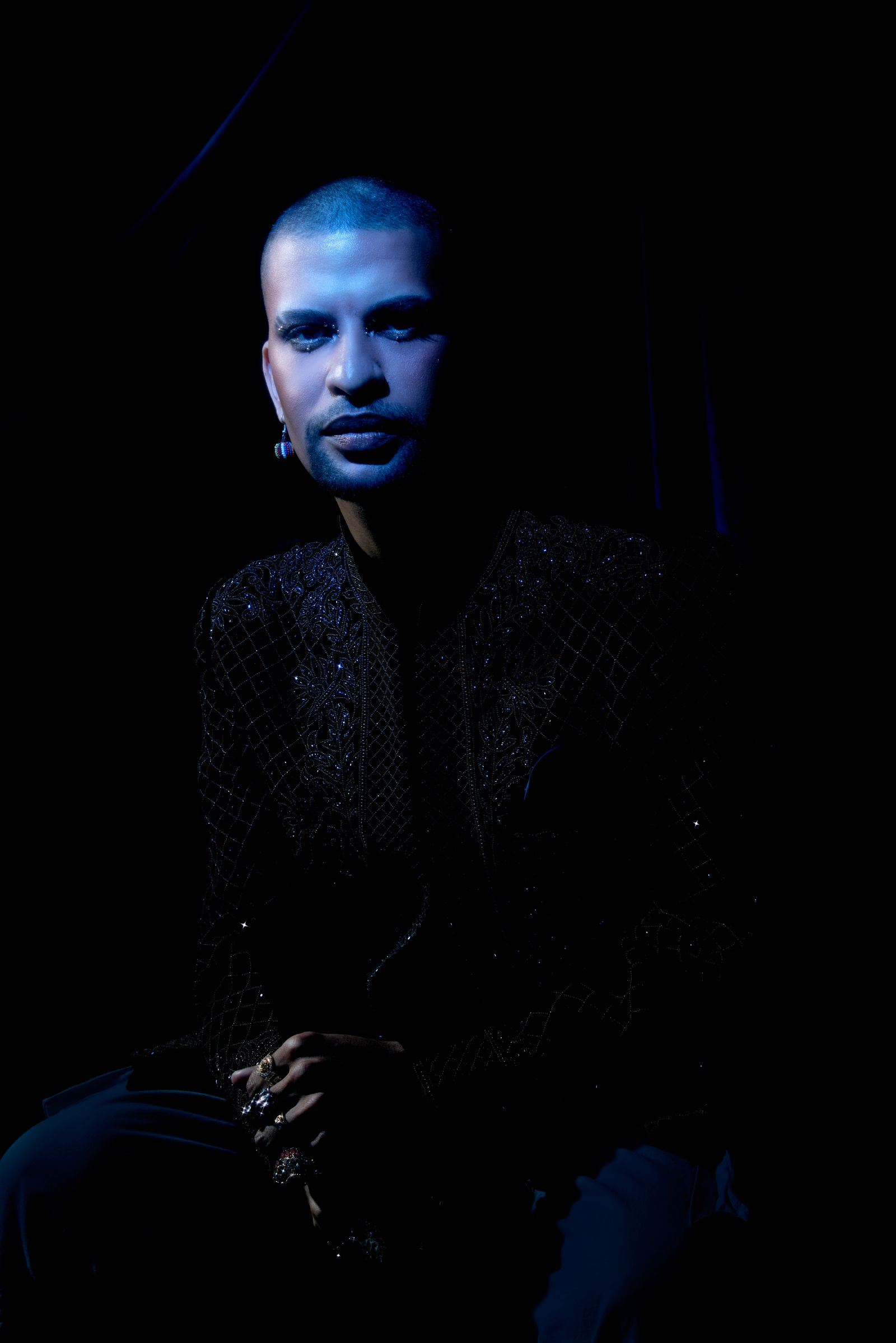 Traidy Naidoo seated, in the same beaded black jacket he wears on the album cover, lit blue against a black ground.