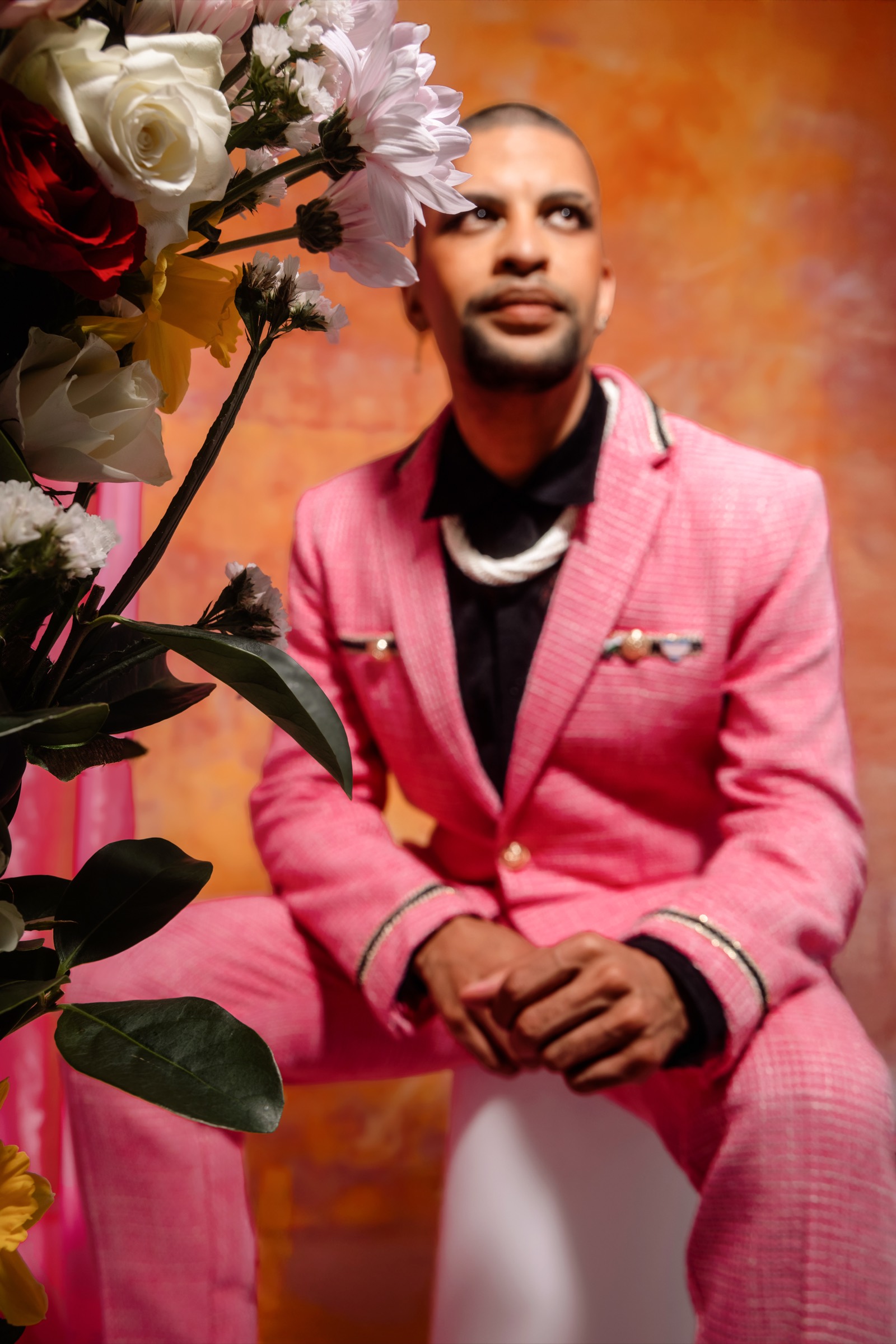 Traidy Naidoo seated in a pink checked suit among roses against an orange backdrop.