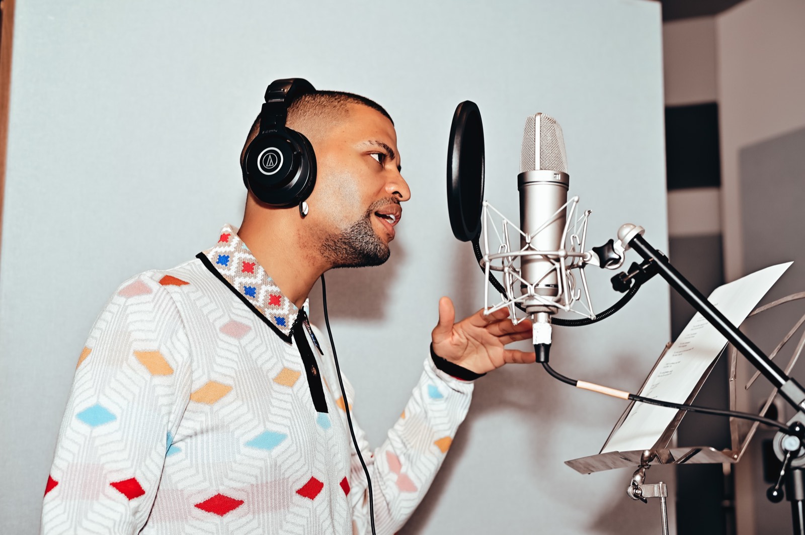 Traidy Naidoo in profile at a studio microphone, headphones on, mid-phrase.
