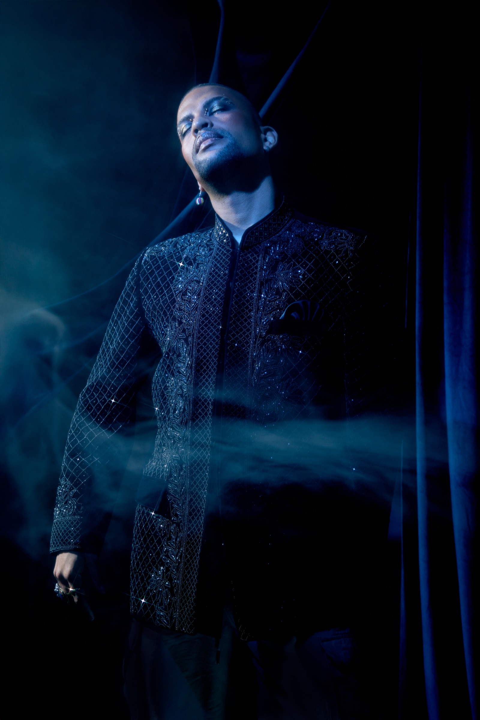 Traidy Naidoo standing in blue light with his eyes closed and his head lifted, smoke drifting across the beaded black jacket.