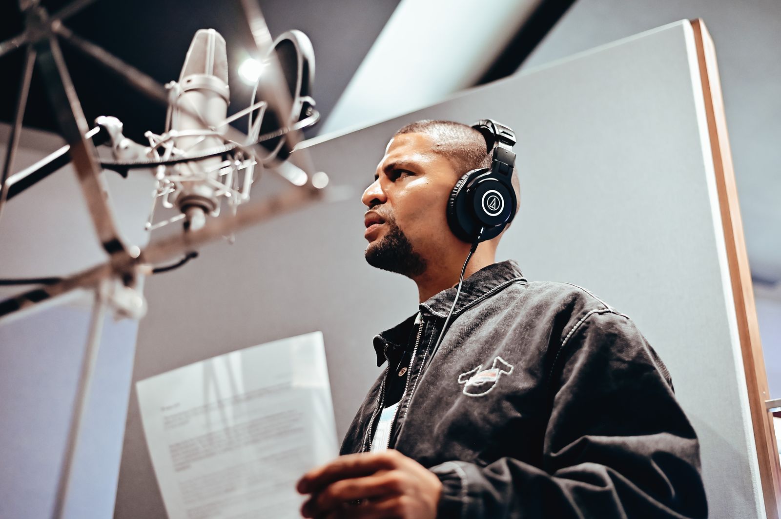 Traidy Naidoo in the recording booth, headphones on, reading from a lyric sheet at the microphone.