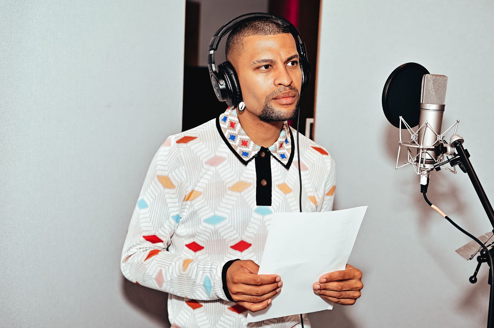 Traidy Naidoo in a recording booth, headphones on, holding a lyric sheet at the microphone.
