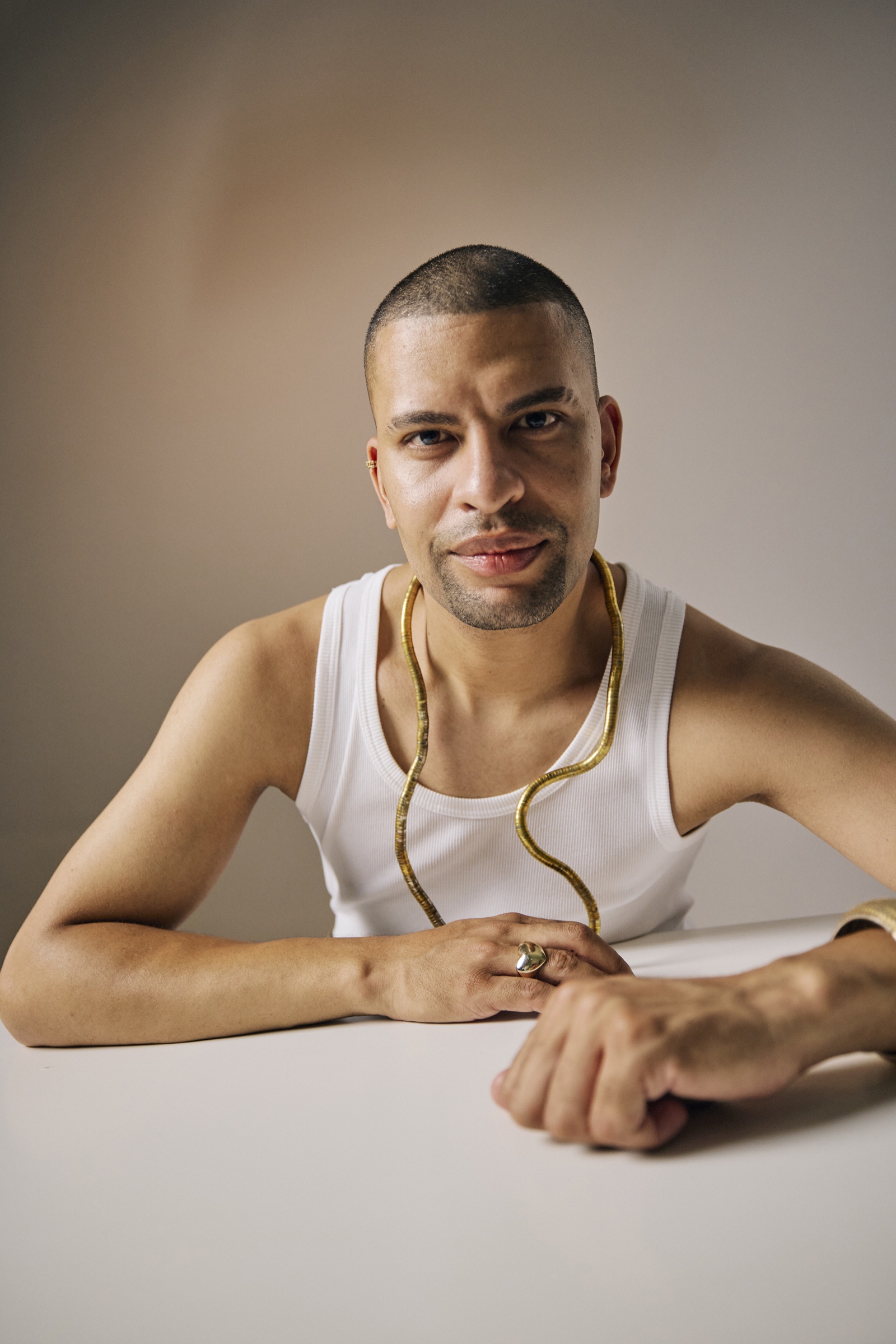 Traidy Naidoo at a white table in a white vest and gold chain, looking to camera.