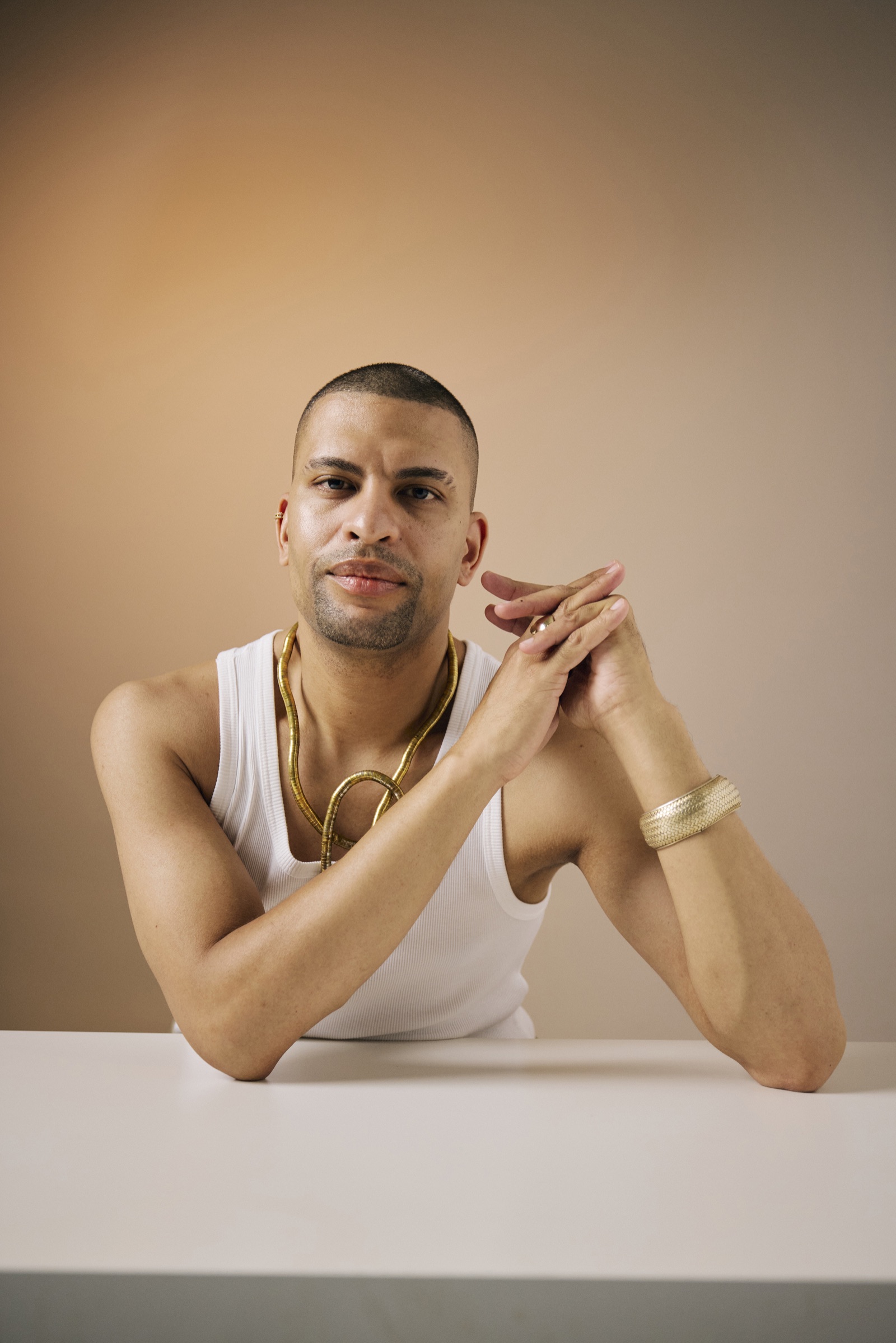 Traidy Naidoo seated at a white table in a white vest, a gold chain and a gold cuff, hands loosely clasped in front of him, looking straight to camera.