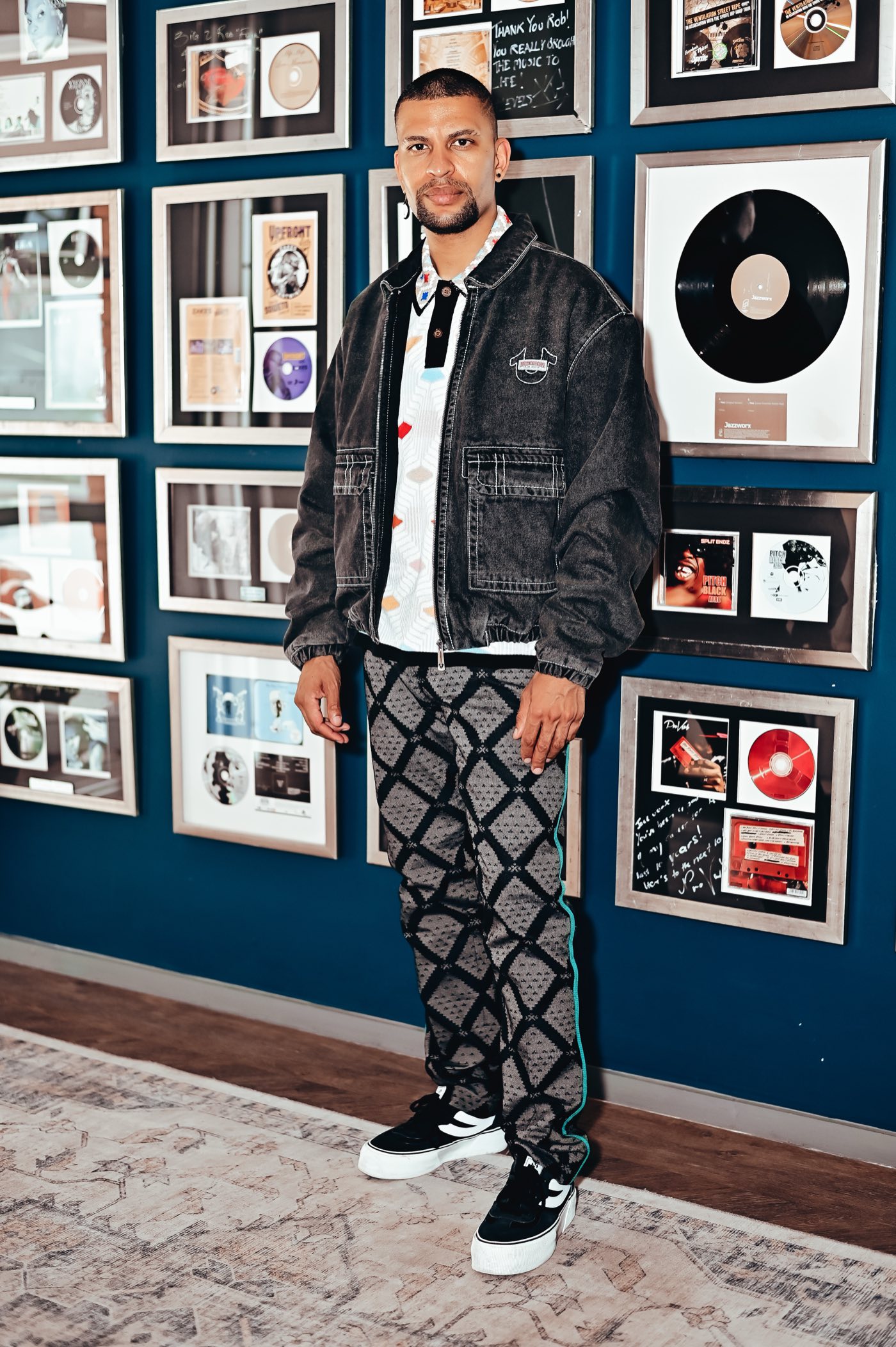 Traidy Naidoo standing in a denim jacket against the studio's wall of framed records.